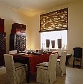 Chairs with slip covers around a table with a red tablecloth in an elegant dining room with bamboo roller blinds