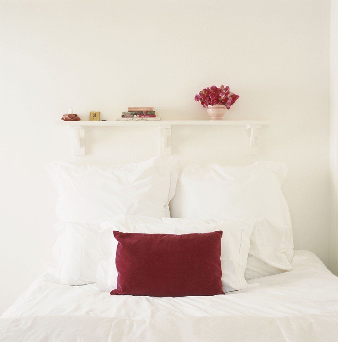 Einzelbett mit weißer Bettwäsche und einem roten Kissen unter kleines Bücherregal