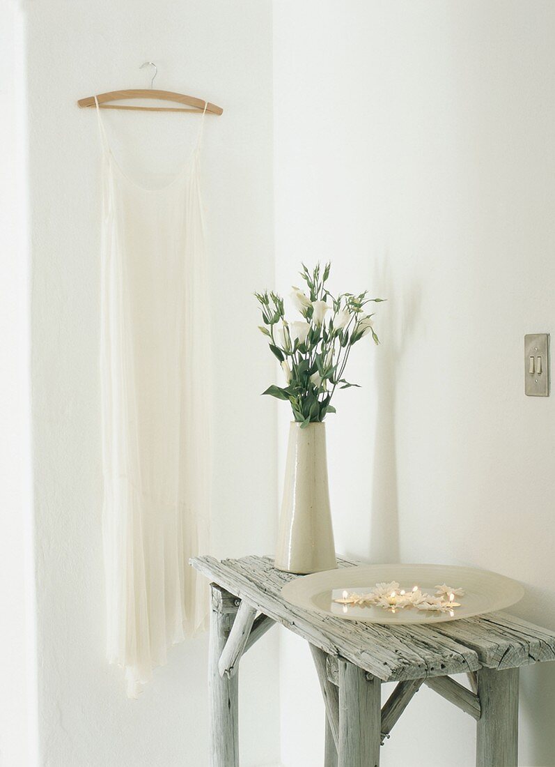 Ein rustikaler Holztisch mit Kerzen und einer Blumenvase vor weißes Damenkleid auf Kleiderbügel