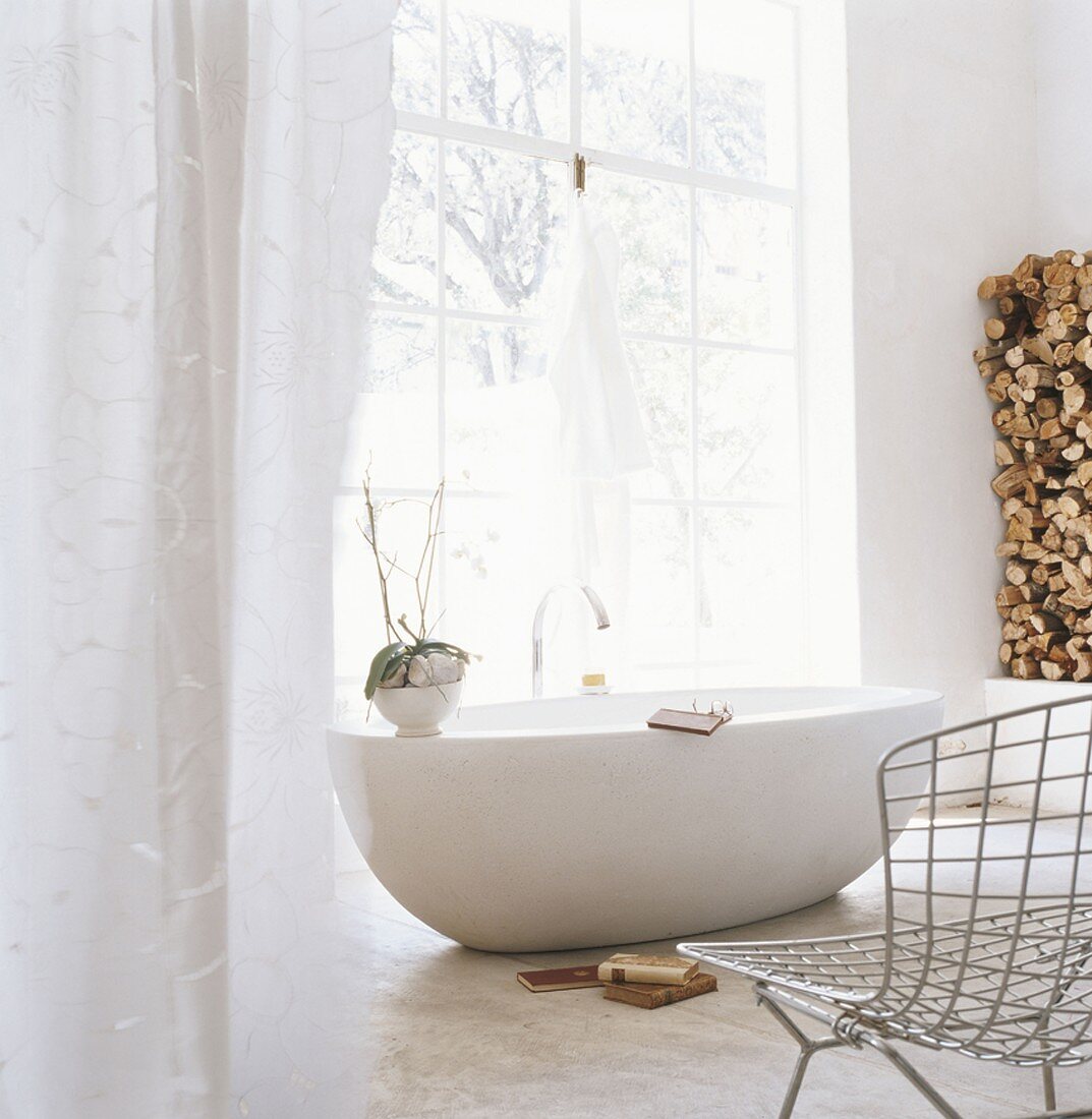 Eine runde freistehende Badewanne vor Panoramafenster im Badezimmer mit Drahtstuhl und gestapletes Brennholz in der Ecke