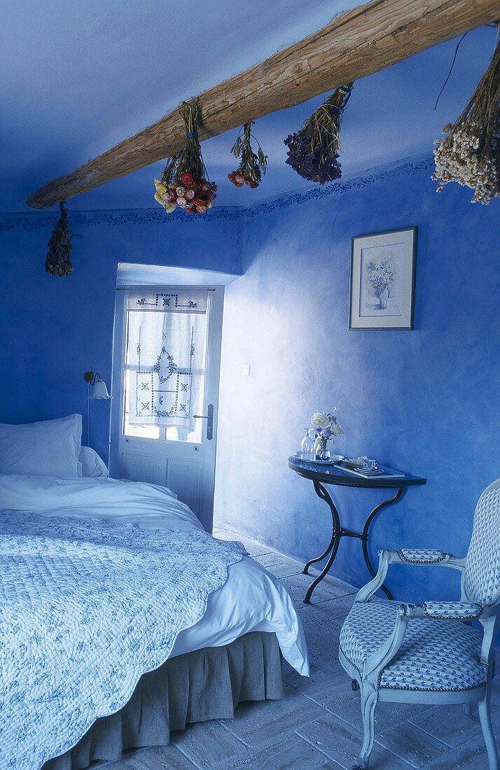 Blaues Schlafzimmer mit Deckenbalken, daran hängenden Trockenblumen, Doppelbett und Steppdecke mit Blümchenmuster