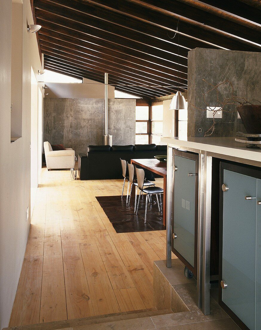Blick von der Küche in einen offenen, modernen Wohnraum mit schräger Balkendecke, Betonwand und einem hellen Holzdielenboden
