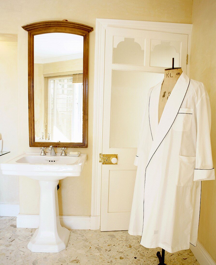 White dressing gown on tailor's dummy in nostalgic bathroom with pedestal sink and large, wood-framed mirror