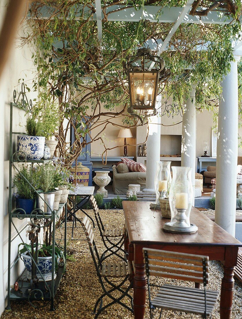 Holztisch mit Klappstühlen auf überdachter Terrasse mit Pergola, Säulen und einem Metallregal mit Blumentöpfen