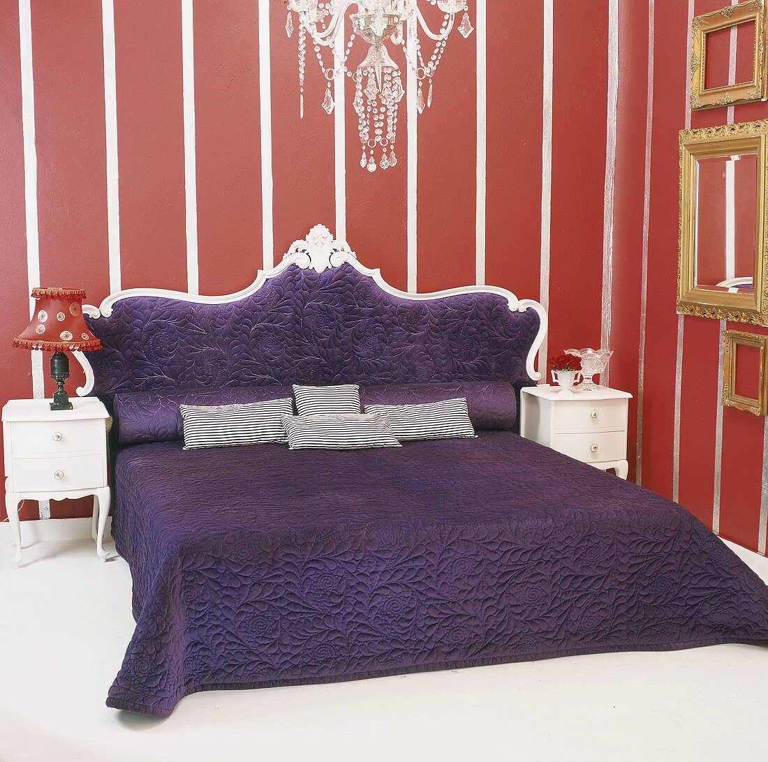 Königliches Doppelbett in Lila mit darüberhängendem Lüster, umrahmt von einer rot-weiss gestreiften Wandtapete und flankiert von einer Sammlung goldener Bilderrahmen