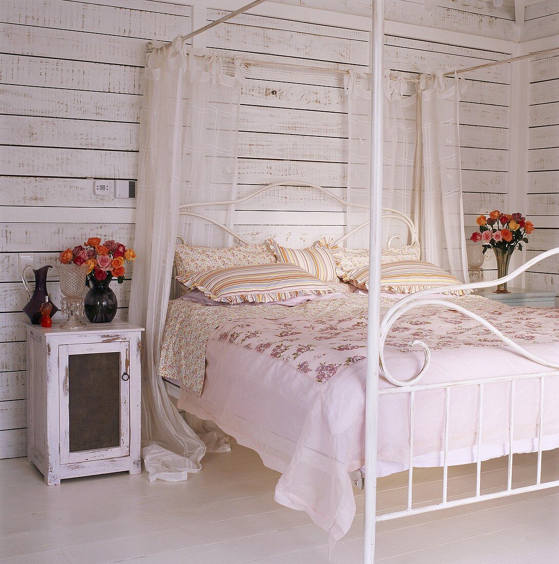 Holztisch mit Rosenstrauß neben schmiedeisernes Himmelbett im Schlafzimmer mit weiß lackierten Holzdielenwänden