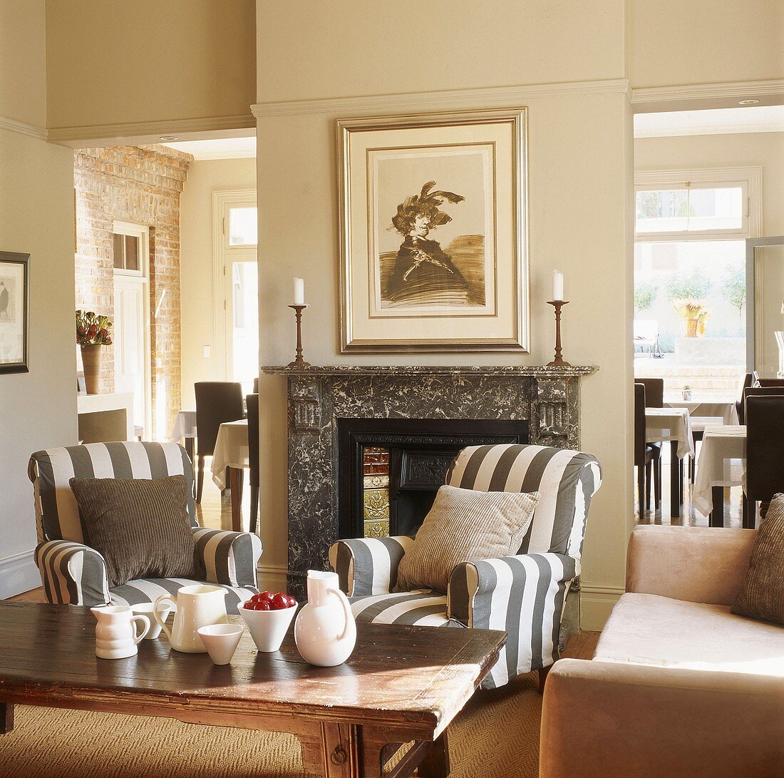 Offenes Wohnzimmer mit gestreiften Sesseln und antikem Couchtisch vor einem Marmorkamin
