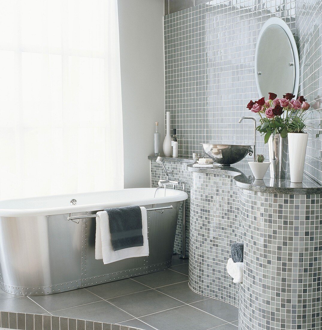Modernes Designerbad mit wellenförmigem Waschtisch aus grauen Mosaikfliesen und freistehende Badewanne in Metall-Optik