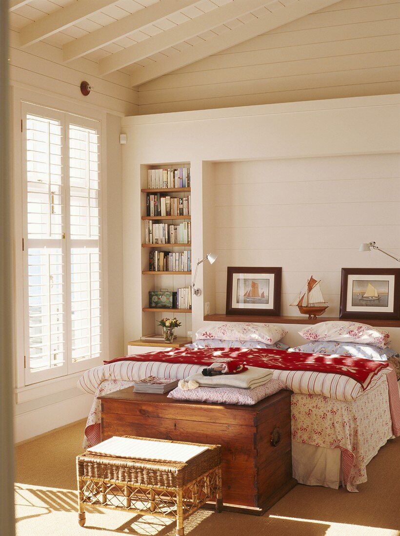 Ein Bett im Mustermix mit antiker Truhe am Fußende in einem hellen Schlafzimmer mit Holzverkleidung