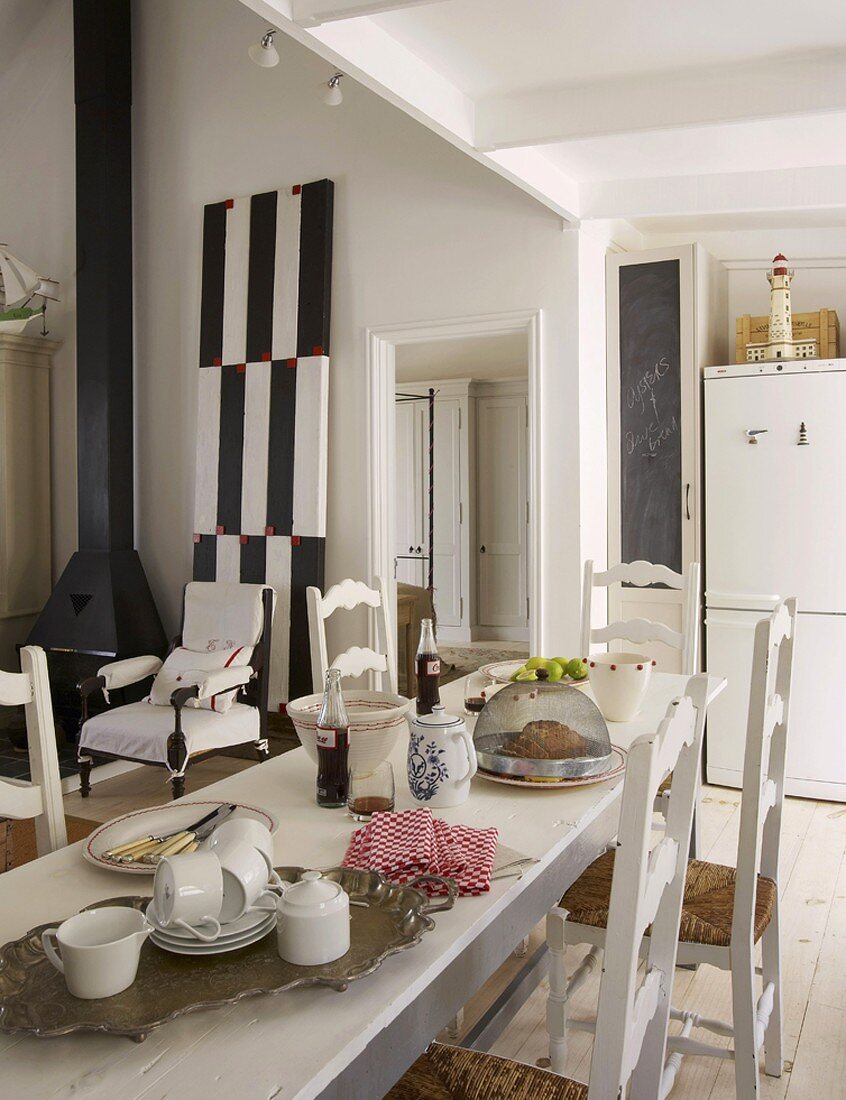 Set, white dining table and white rush-bottom chairs; black wood-burner and black and white artwork in background