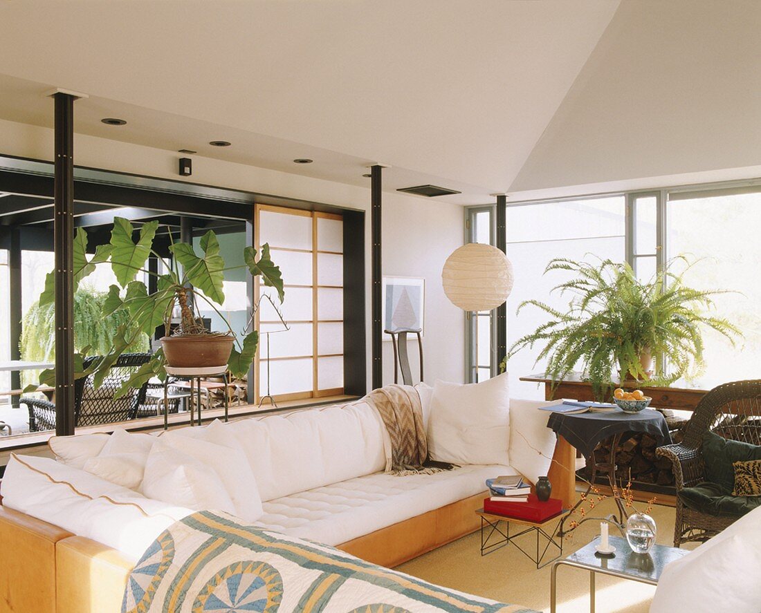 Wohnzimmer mit großer, weisser Eckcouch; dahinter eine überdachte Terrasse mit einem Korbstuhl und eine exotische Zimmerpflanze