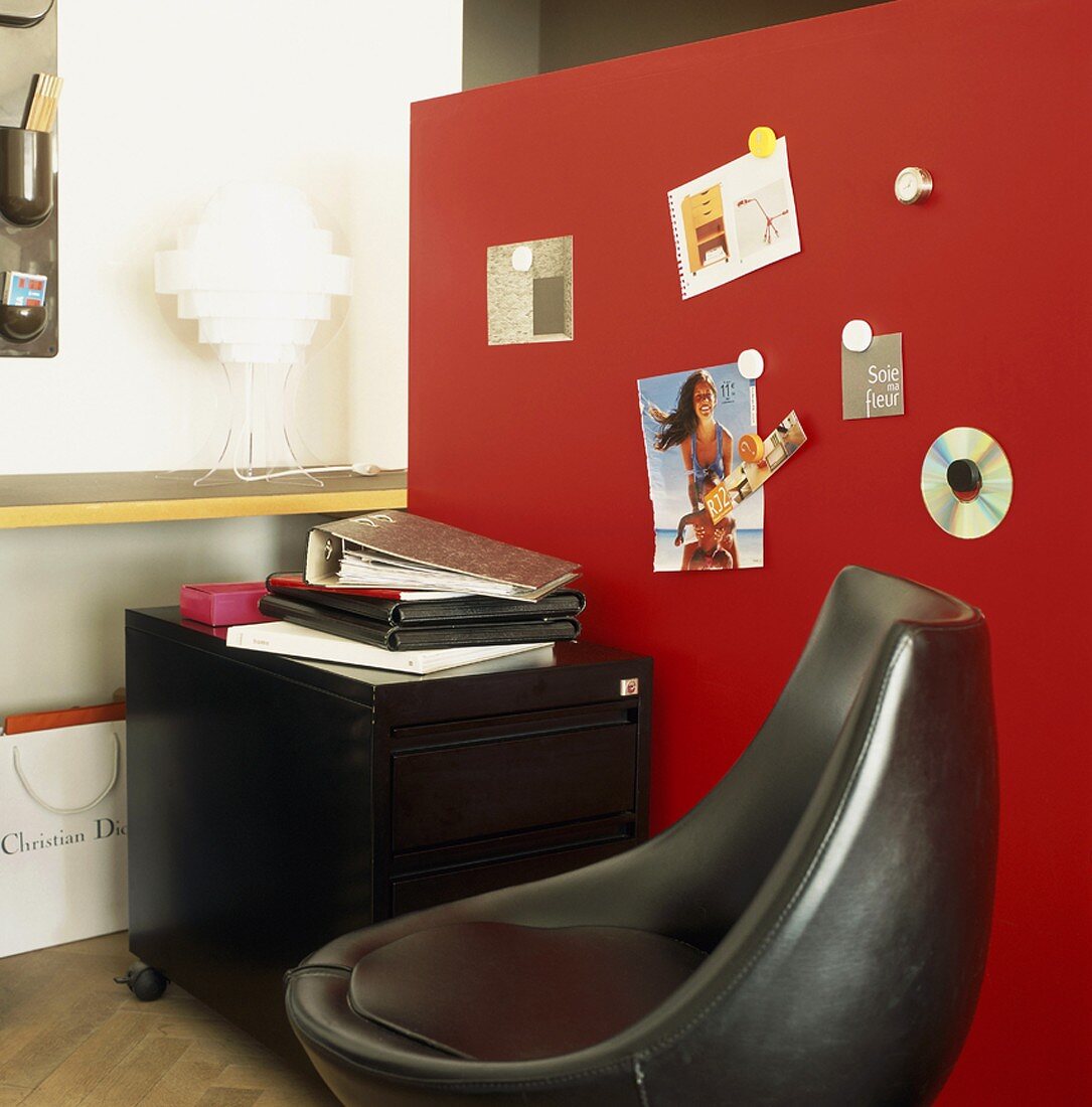 Arbeitsecke mit schwarzem Rollcontainer vor einer roten Pinwand; davor ein schwarzer Designersessel