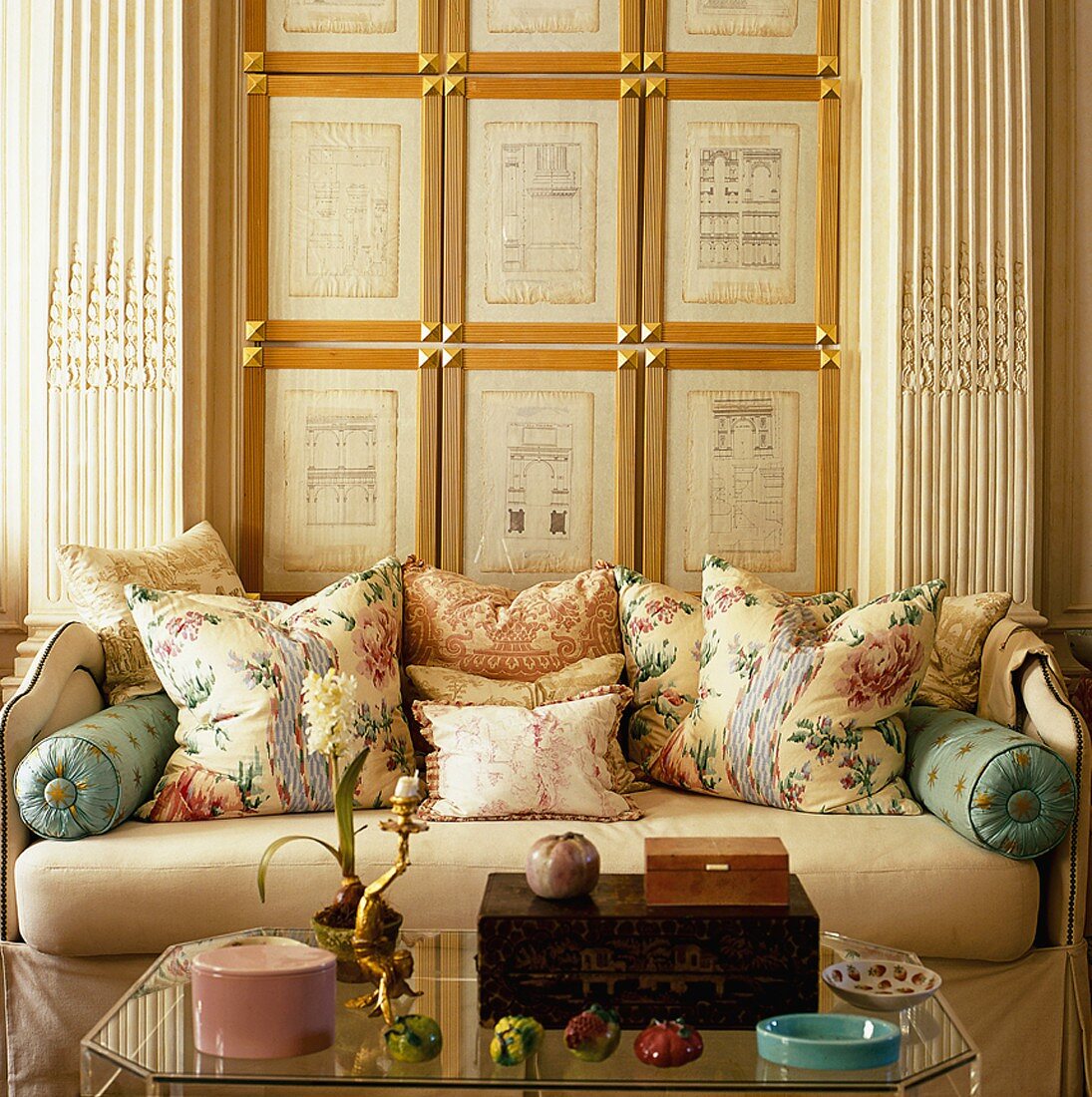 Gerahmte Architekturskizzen zwischen Pilastern, davor ein Sofa mit verschiedenen Kissen und ein gläserner Couchtisch