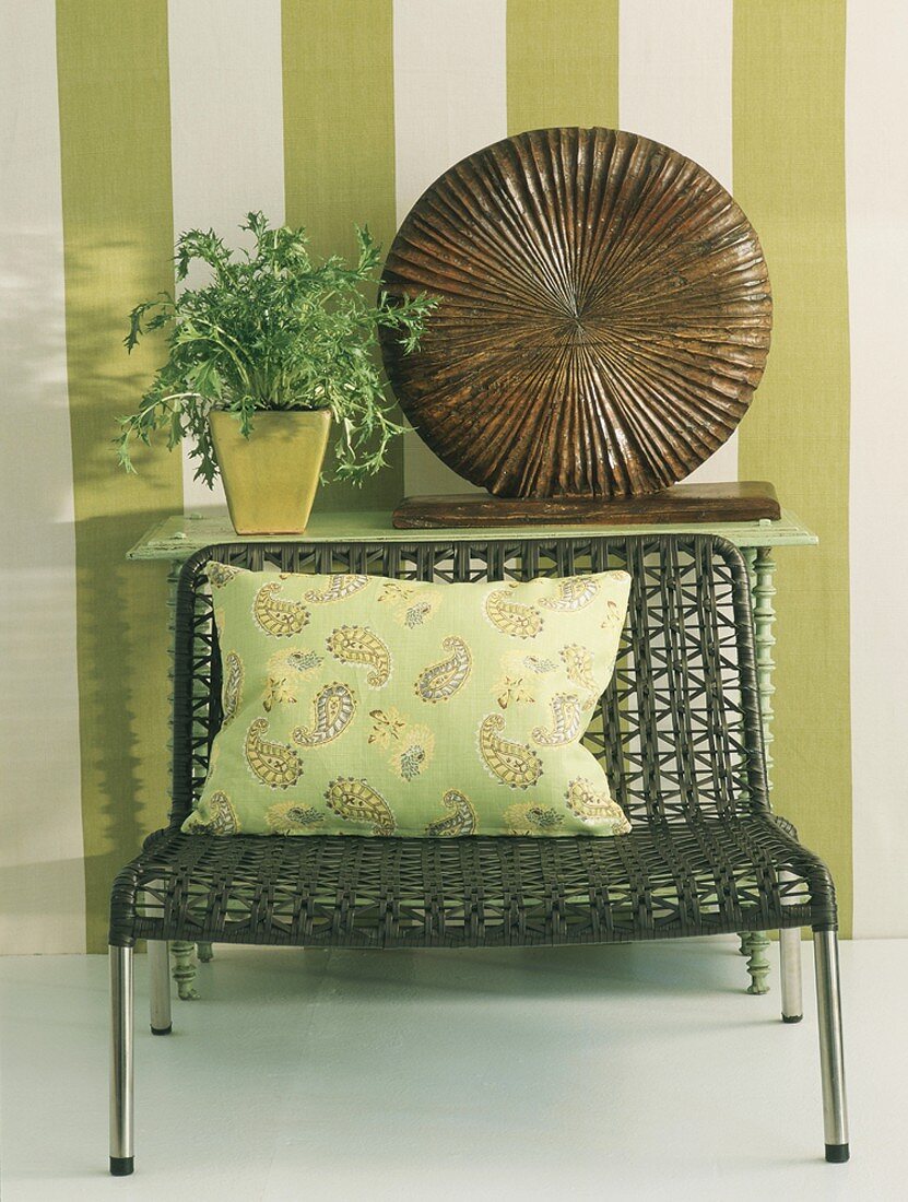 Geflochtener Stuhl mit Kissen vor einem Wandtisch mit Holzskulptur und Blumenvase