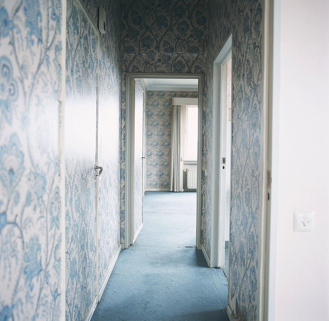Blick in den Flur einer leerstehenden Wohnung mit Barocktapete und blauem Teppichboden