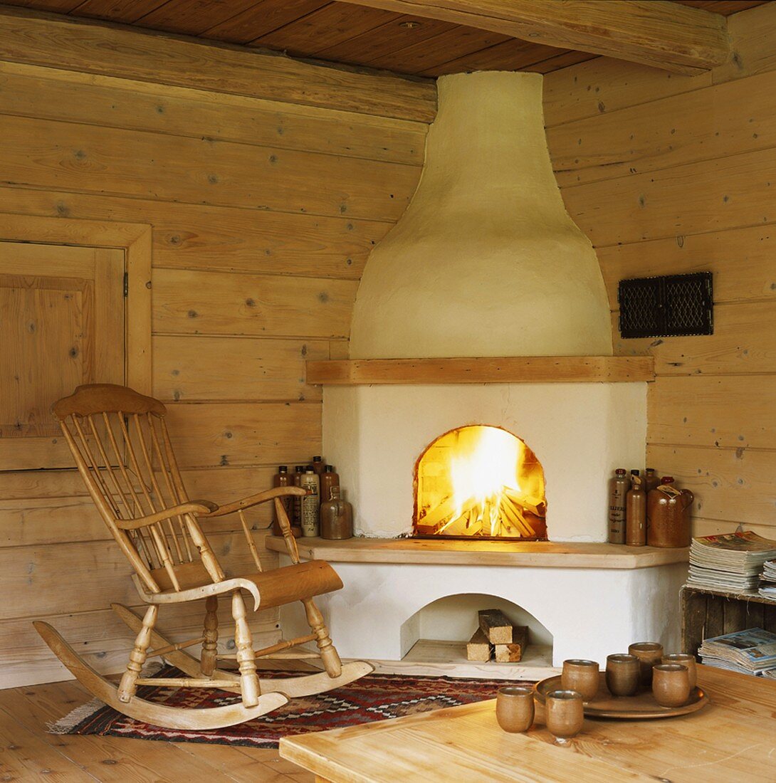 Ein traditioneller Eckkamin aus Lehm und ein Schaukelstuhl in einer urigen Holzhütte