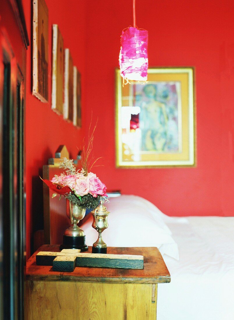 Ein rot gestrichenes Schlafzimmer mit kleinen Pokalen und Holzkreuz auf einem antiken Beistelltisch