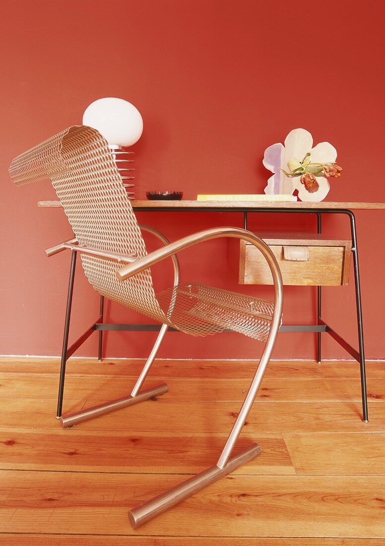 Designerstuhl mit Sitzfläche aus Lochblech am schlichten Vintageschreibtisch vor einer roten Wand