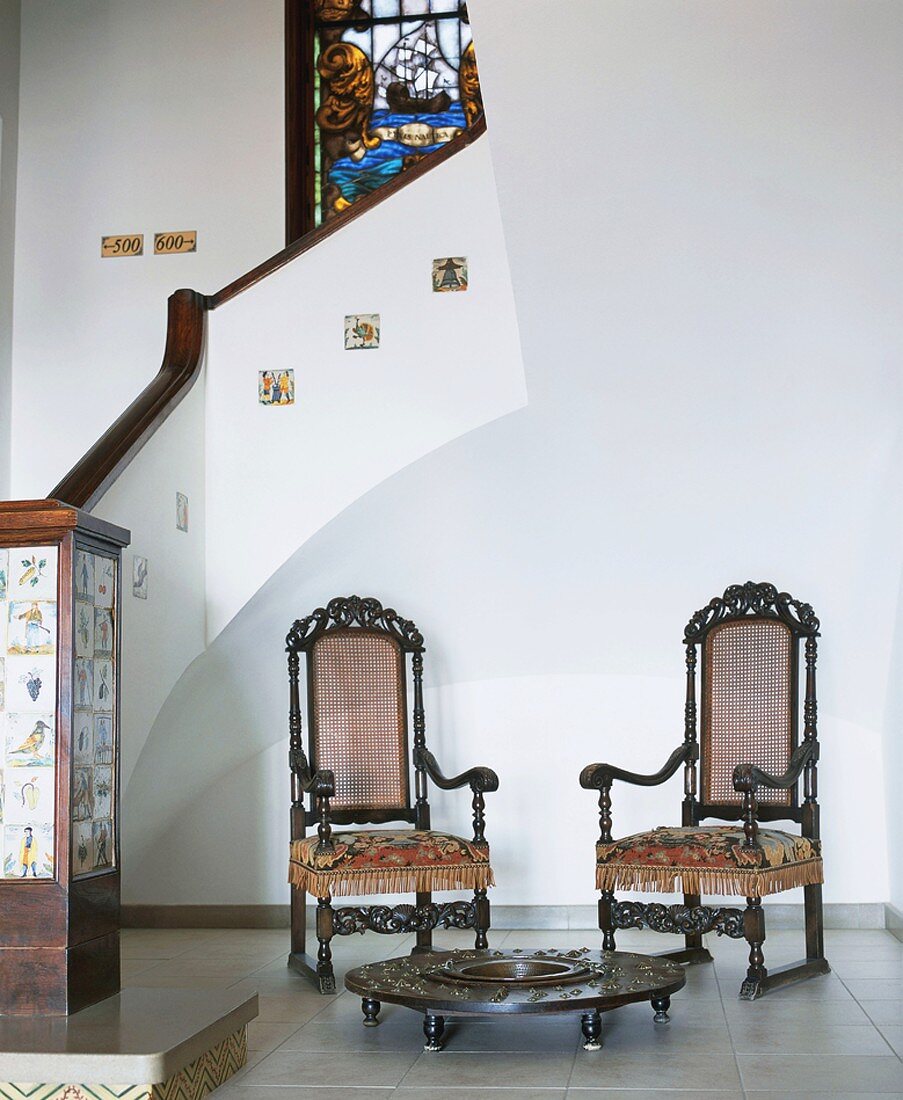 Zwei antike Stühle mit Bodentisch am Absatz eines historischen Treppenhauses mit Mosaikdetails und Fenstermalerei