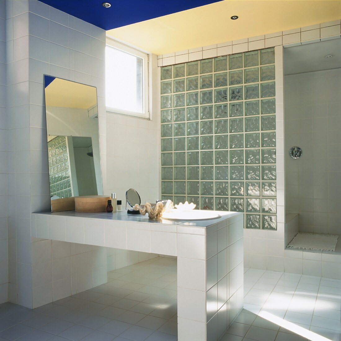 Eine Wand aus Glasbausteinen trennt die Dusche vom weiss gefliesten Badezimmer mit gemauertem Waschtisch ab