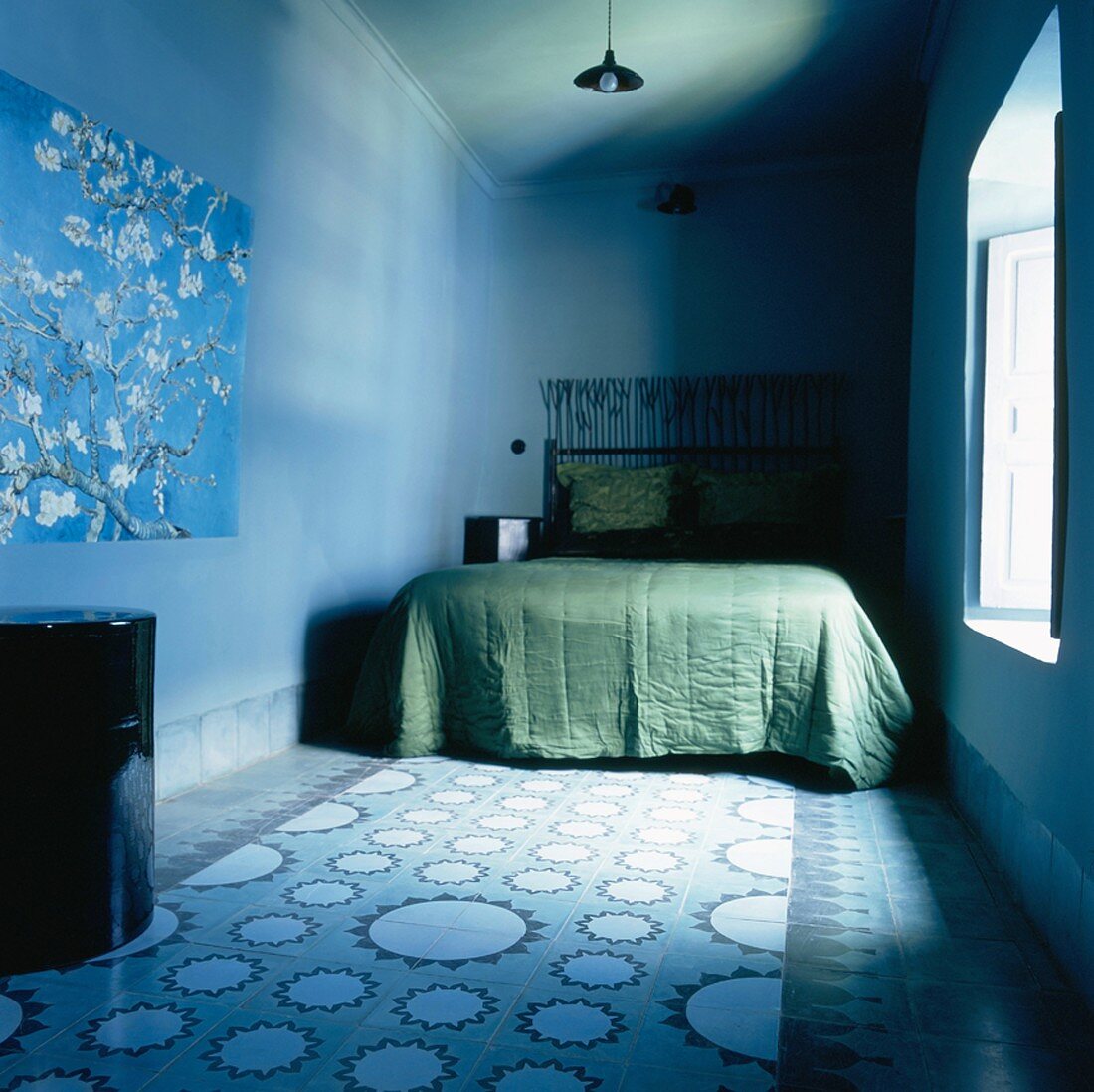 Ein blau gehaltener, einfacher Schlafraum mit grünem Bett und gemusterten Bodenfliesen