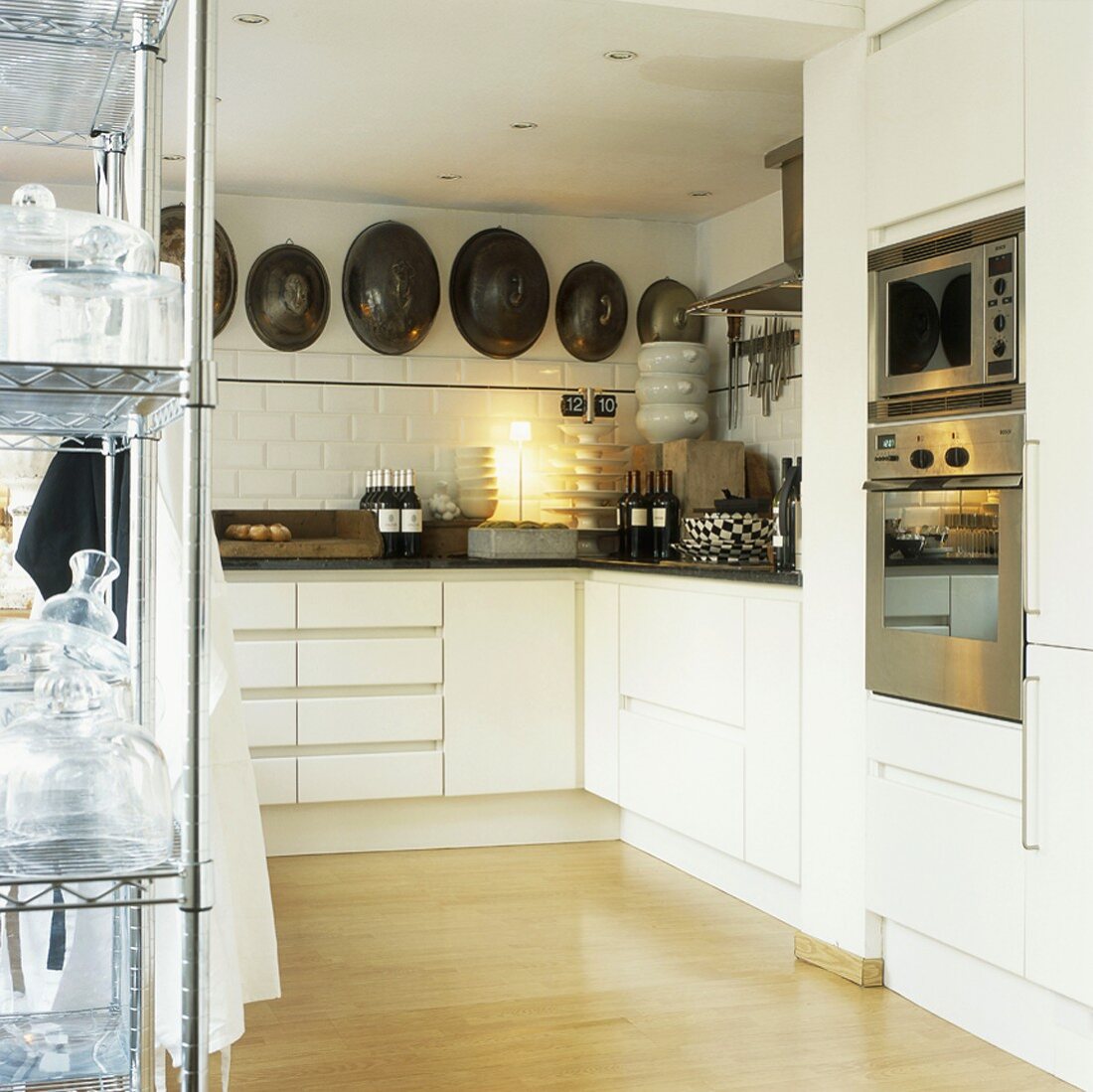 Antike Servierglocken zieren die Steinwand der weißen, minimalistischen Küchenzeile