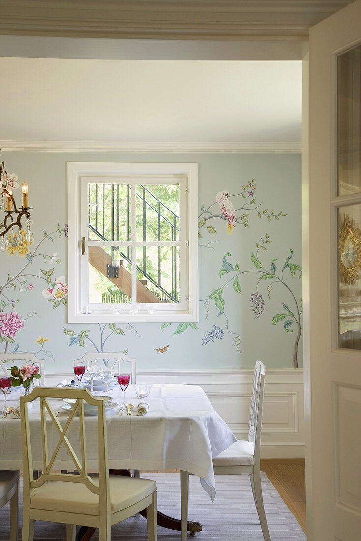 Blick auf einen gedeckten Esstisch vor einer Wand mit Sprossenfenster und floraler Wandmalerei