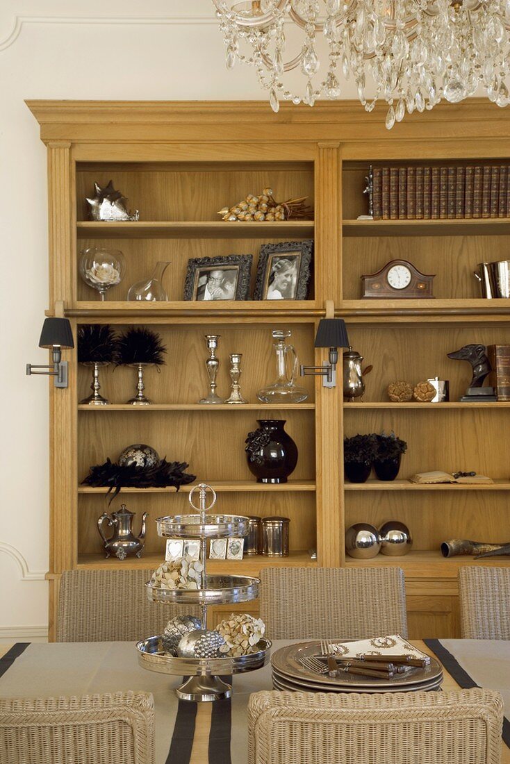 Diverse Wohnaccessoires in einem Schrankregal hinter einem Esstisch mit Silberetagère und Rattanstühlen