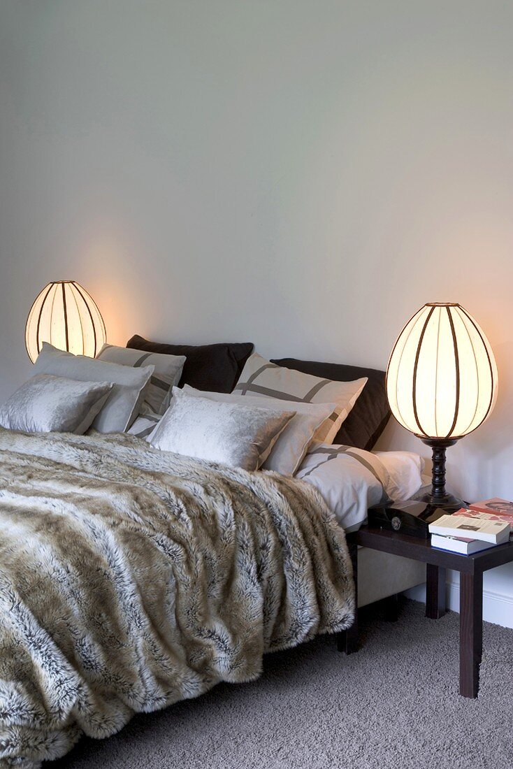 Silberne Kissen auf Doppelbett mit Felldecke und ellipsenförmigen Nachttischlampen