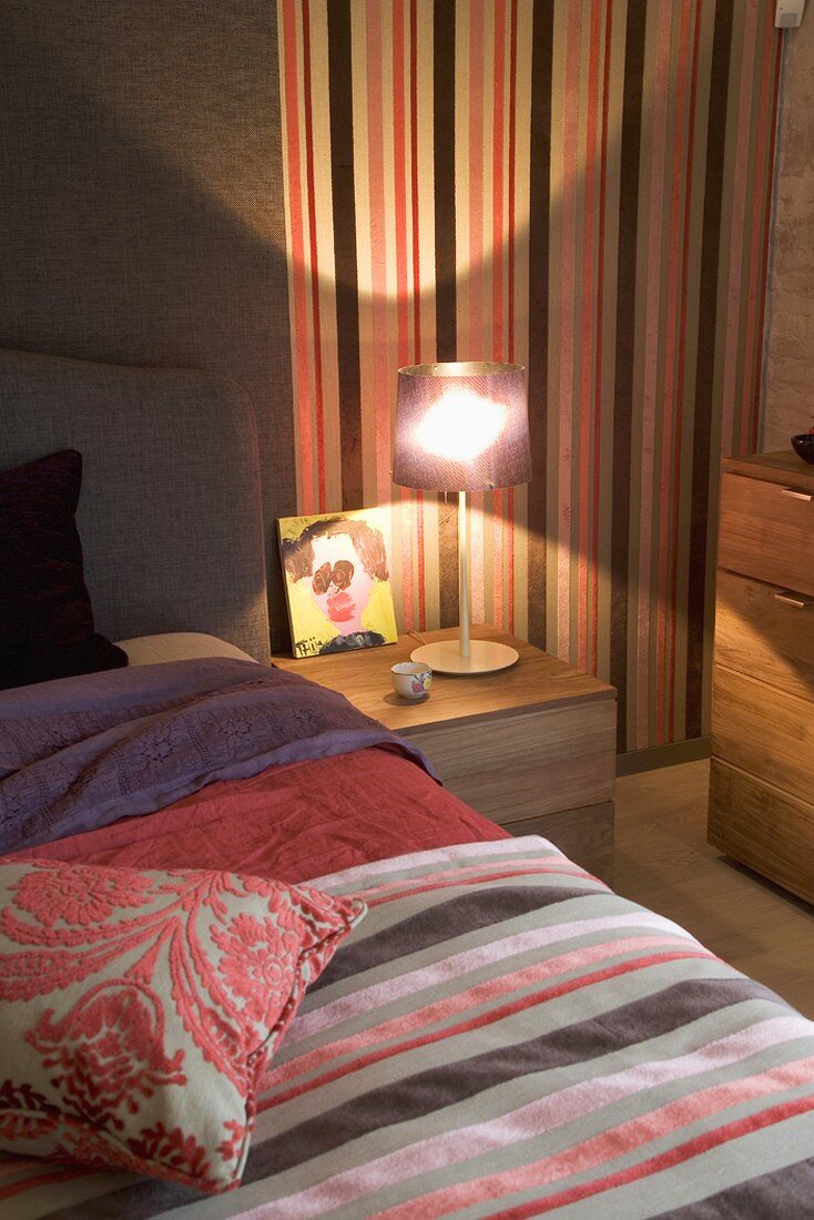 Ausschnitt eines beleuchteten Schlafraums mit Bett, Nachttischlampe, gestreifter Wandtapete & passender Tagesdecke
