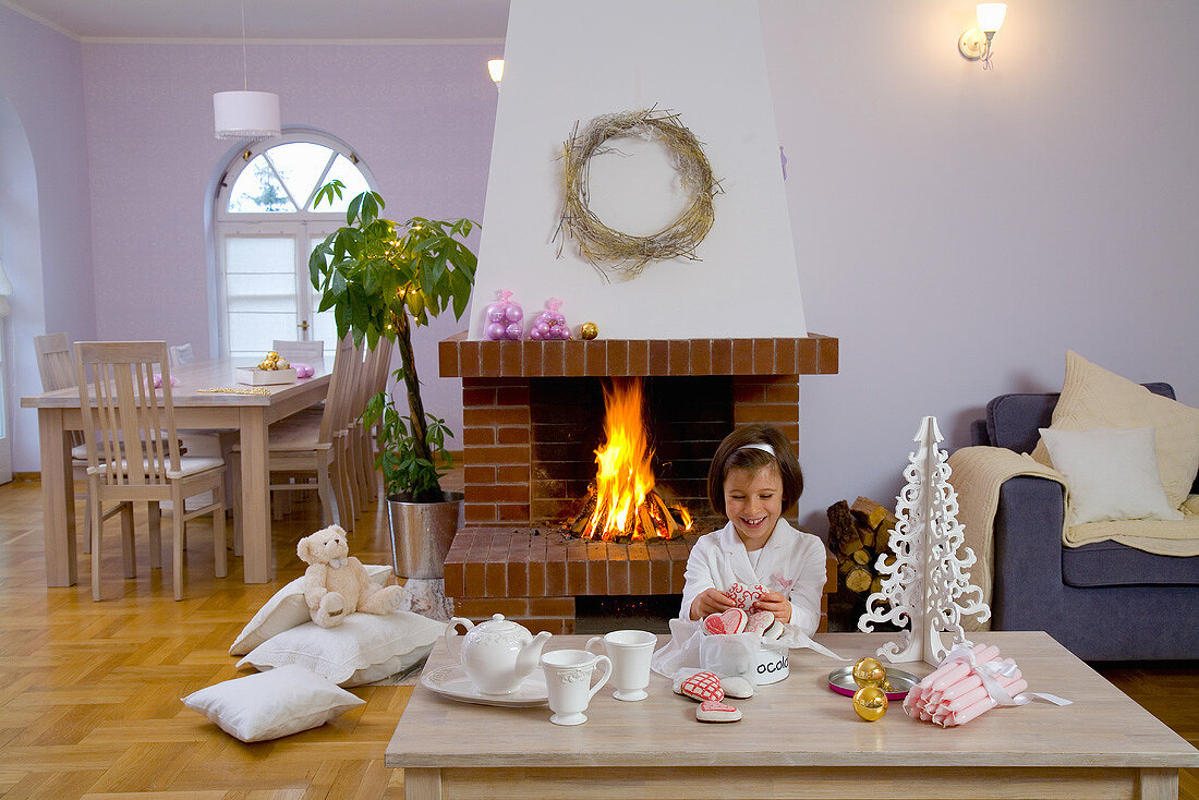 Mädchen am Tisch vor offenem Kamin in weihnachtlich dekoriertem Wohnzimmer