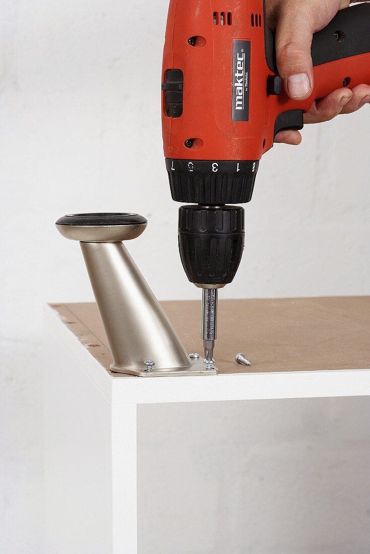 Metal feet being attached to a cupboard using an electric screwdriver