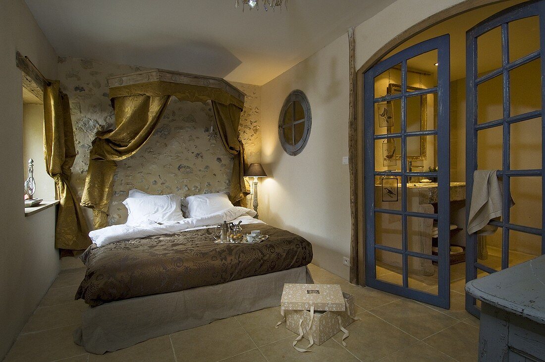 Schlafzimmer mit Doppelbett, Betthimmel aus goldfarbenen Vorhängen & geöffneter Glastür zum Bad