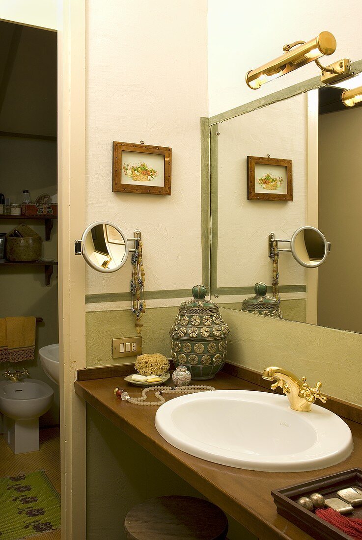 Corner of a bathroom with washstand and brass fittings in front of a mirror and open door with a view into a toilet