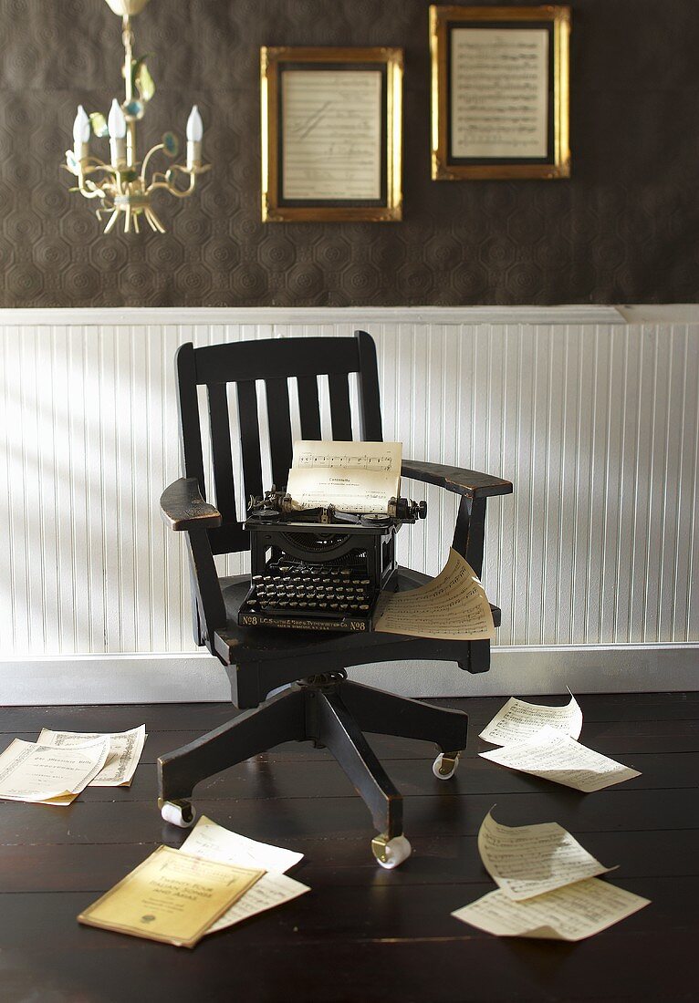 Alte Schreibmaschine auf schwarzem Drehstuhl, rundherum verstreute Notenblätter
