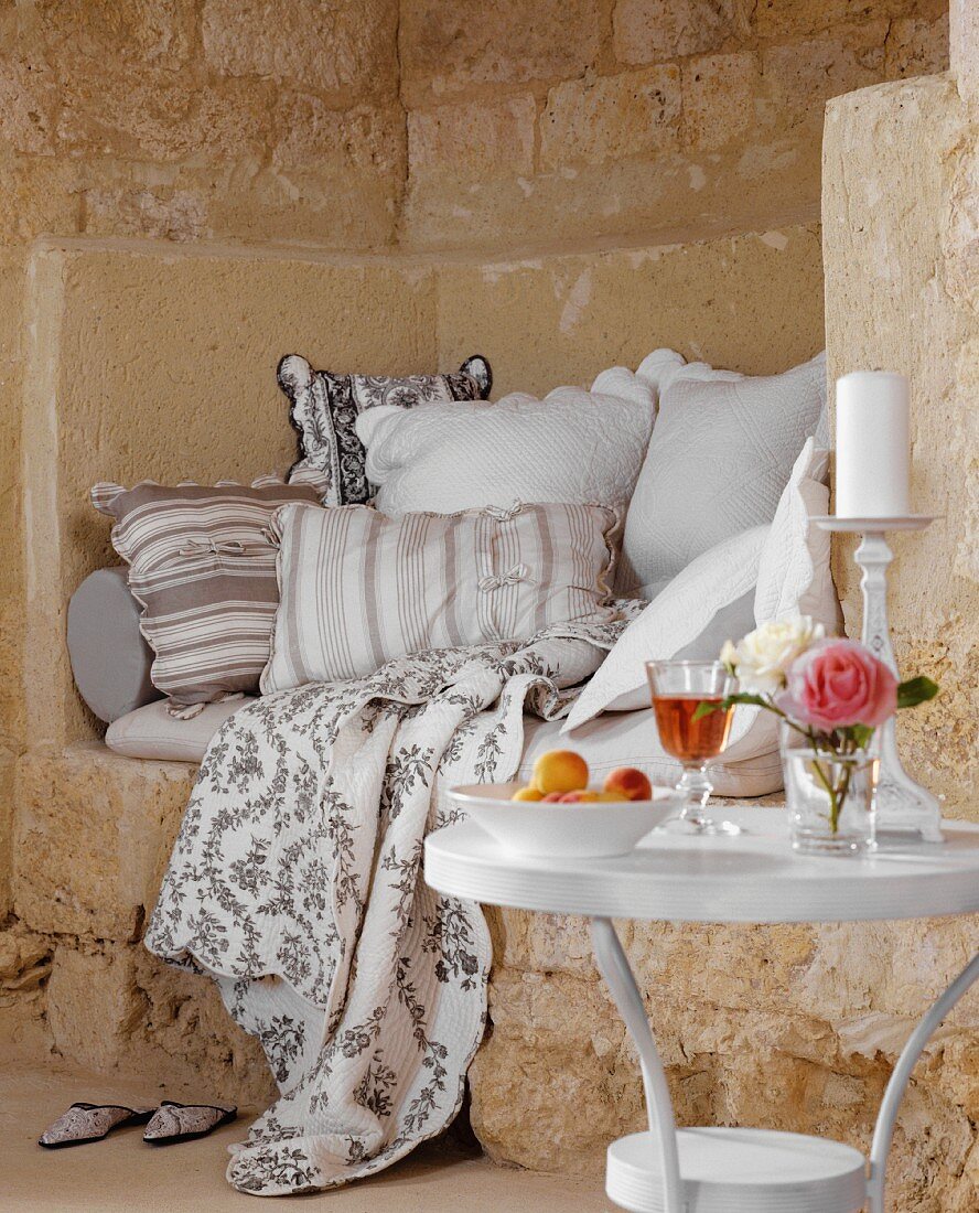 Rustikale Sitznische mit weichen Kissen und Sitzpolster; davor ein kleiner Tisch mit Wein, Obst und einer Rose
