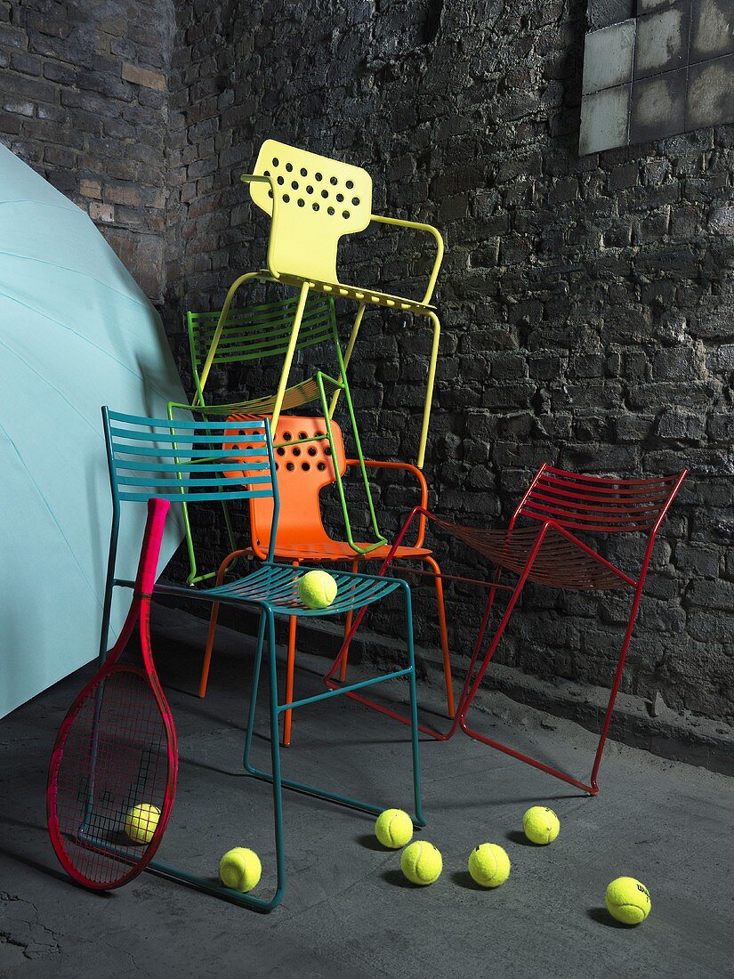 Wildgestapelte Metallstühle in verschiedenen Farben vor einer grauen Steinfassade; am Boden rollende Tennisbälle