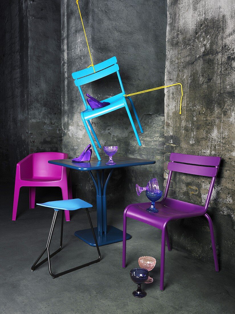Mit Seilen aufgehängter, azurblauer Stuhl, darunter ein dunkelblauer Bistrotisch und violette Plastikstühle; dahinter eine graue, verwitterte Wand