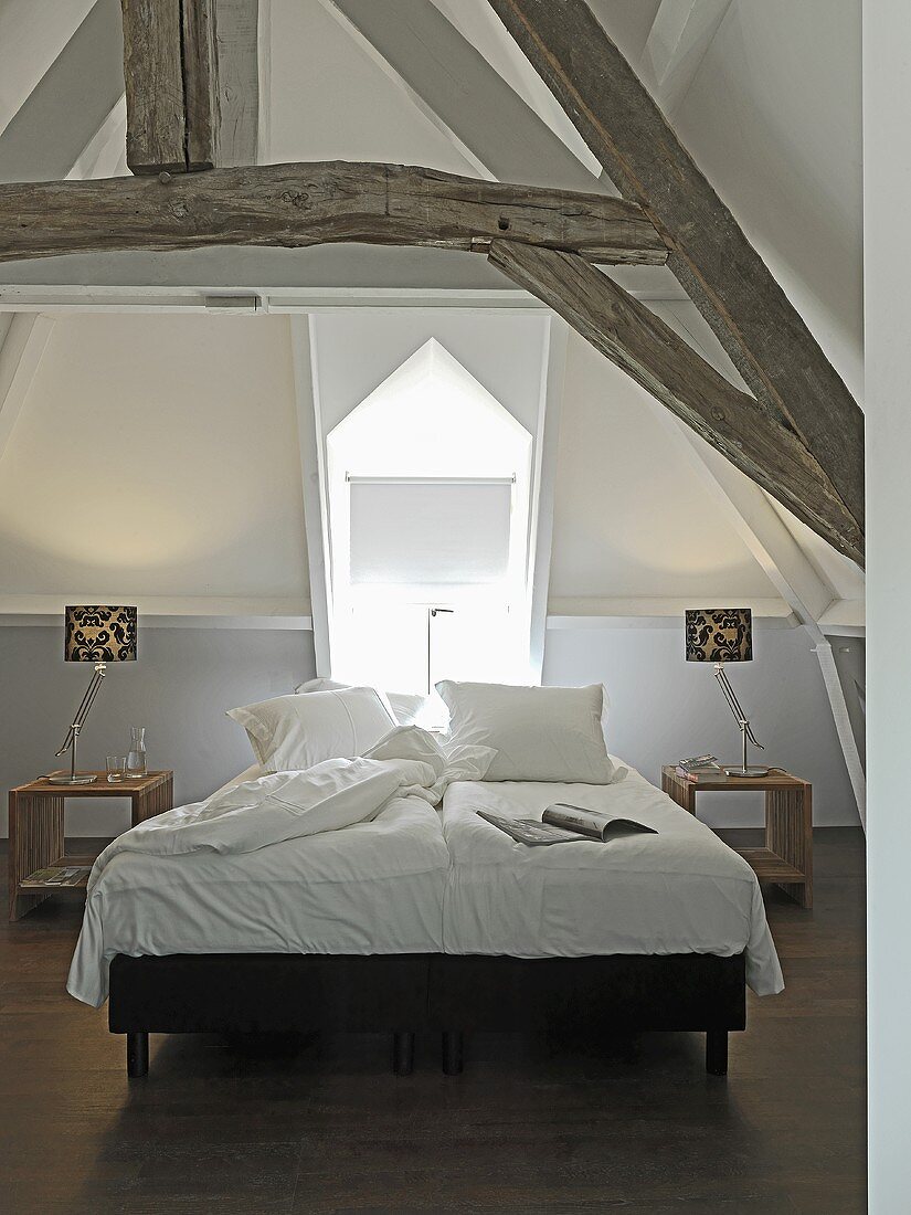 Weisser Schlafraum mit Doppelbett im ausgebautem Dach mit sichtbarer, rustikaler Holzkonstruktion
