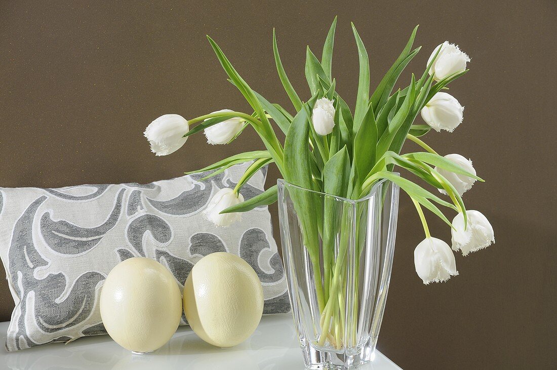 A display featuring white tulips in a glass vase, ostrich egg and a cushion