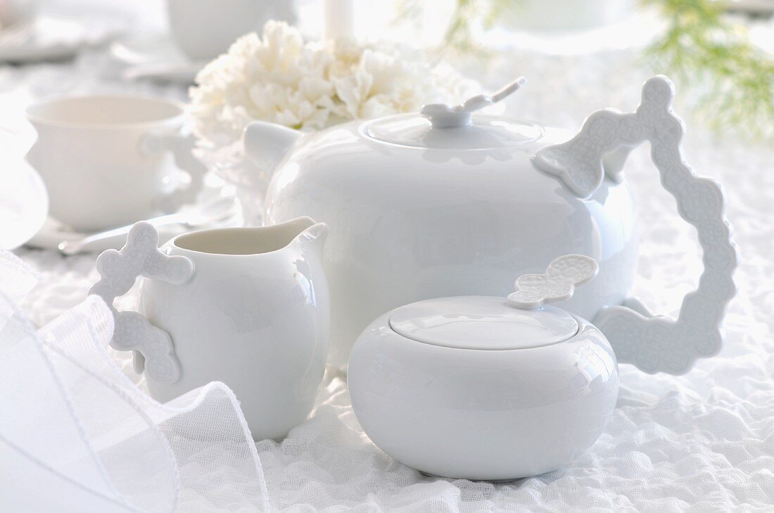 weiße Teekanne mit Zuckerdose und Milchkännchen auf weisser Tischdecke