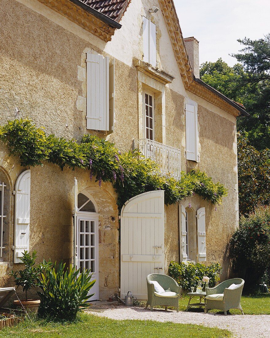 Fassade eines alten Landhauses mit Fensterläden & Rattanstühlen auf Kiesweg (Frankreich)