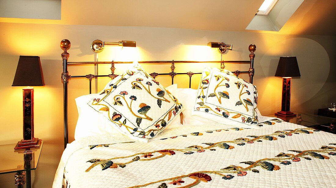 Beleuchtetes Schlafzimmer mit Doppelbett aus Metall, Tagesdecke & Zierkissen mit Blumenmotiv, Wandleuchten & Tischleuchten auf Glasnachttischchen