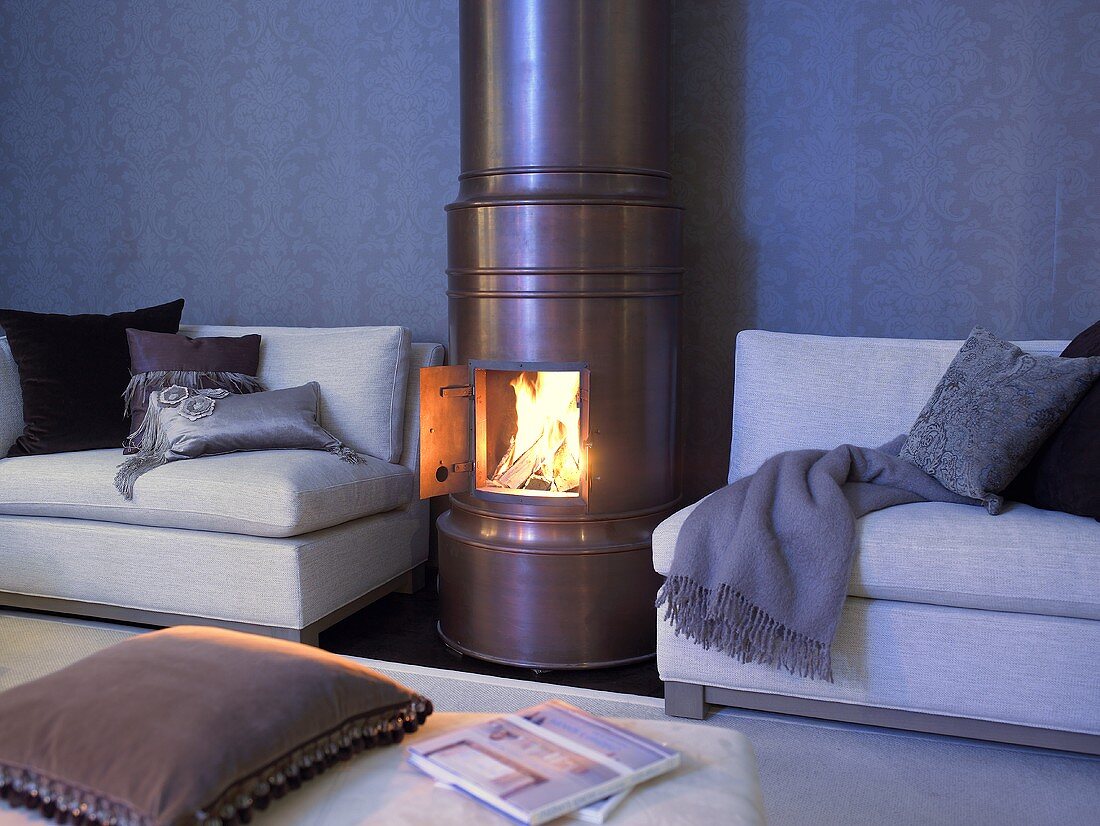Kupferfarbener Kaminofen mit offener Tür und Blick auf Feuer neben Sofa mit Kissen und Decke