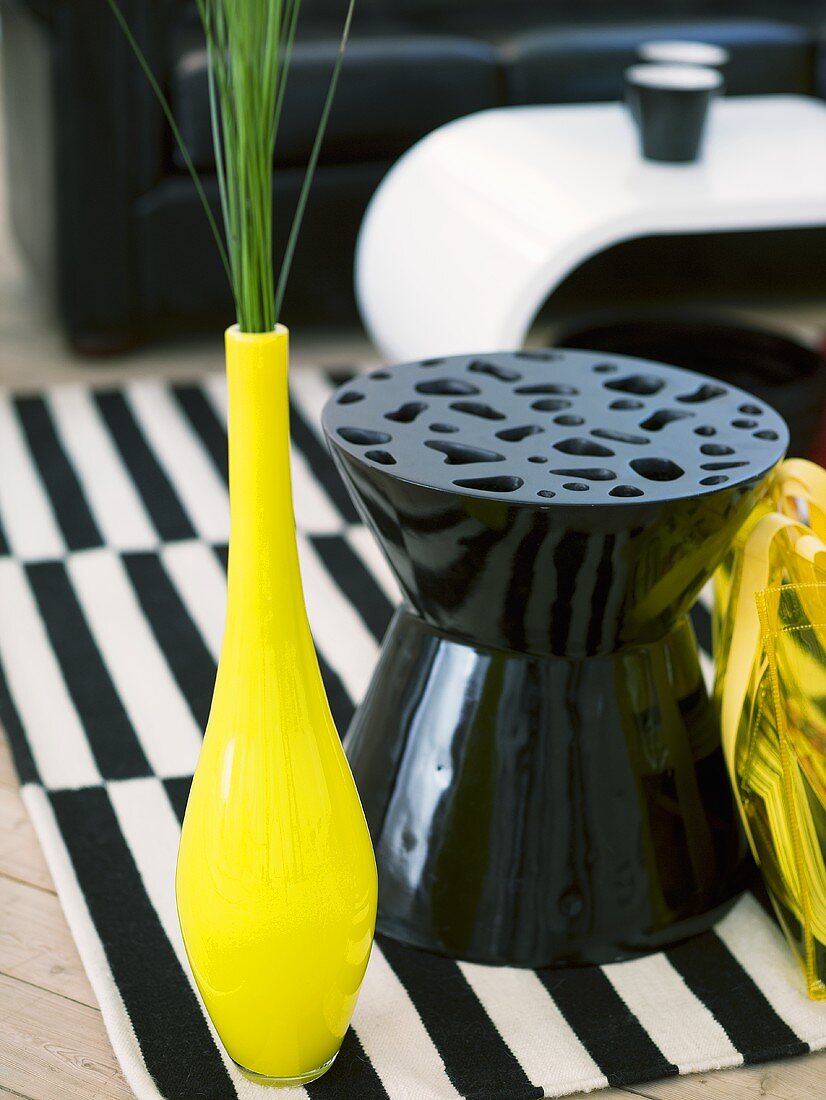 Yellow vase with ornamental grass next to a black plastic stool on a black and white carpet