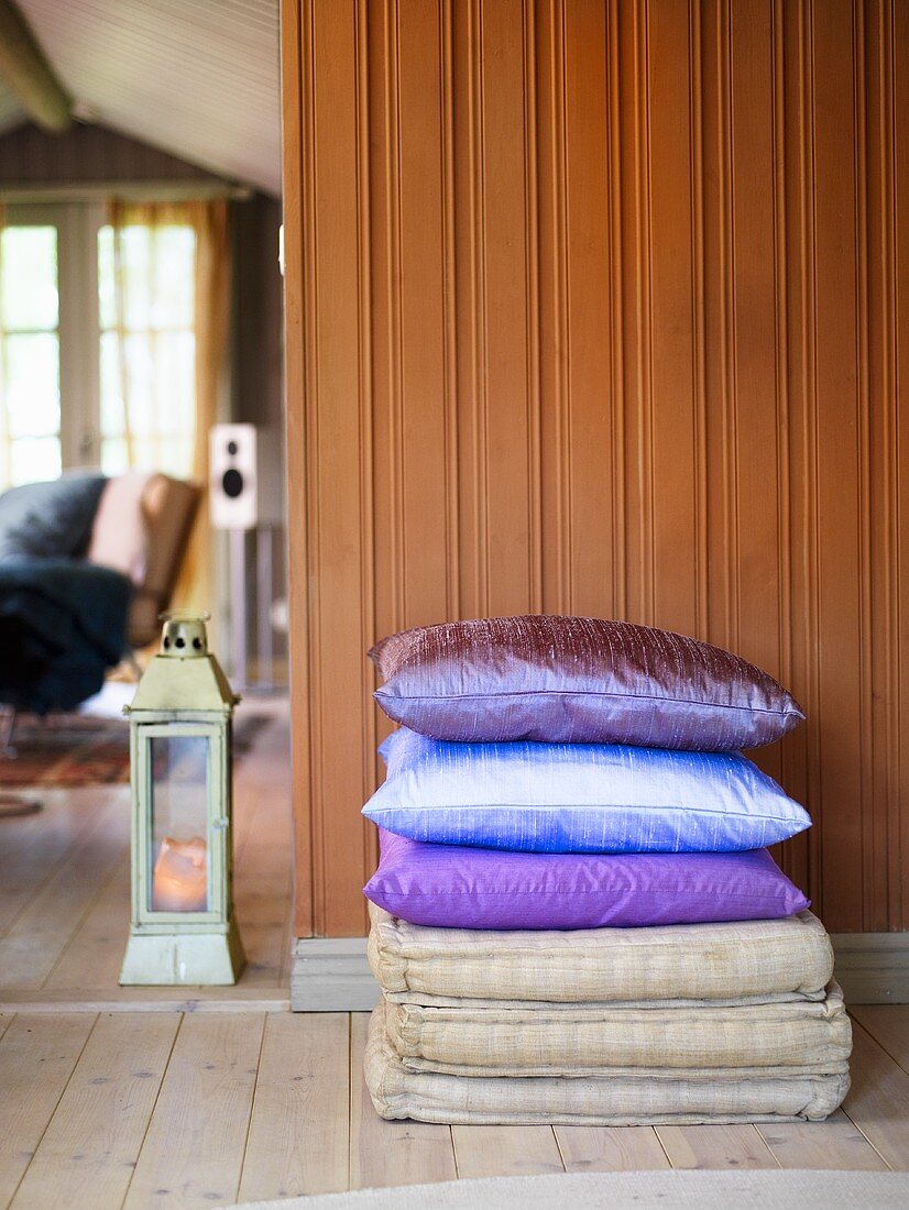 Kissenstapel mit glänzendem Seidenbezug vor brauner Holzwand neben Durchgang mit Blick in Wohnraum