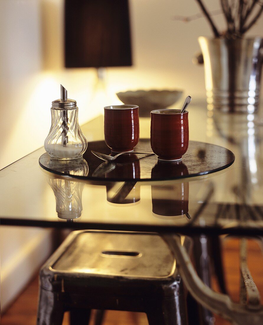 Zwei Trinkbecher & ein Zuckerstreuer auf Glastisch in Wohnraum