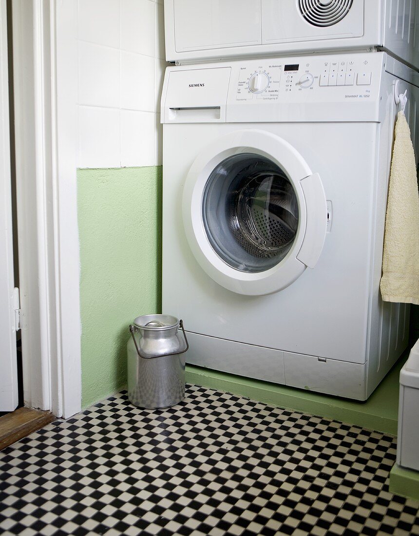 An old milk can in front of a washing machine in a bathroom with a chessboard pattern floor