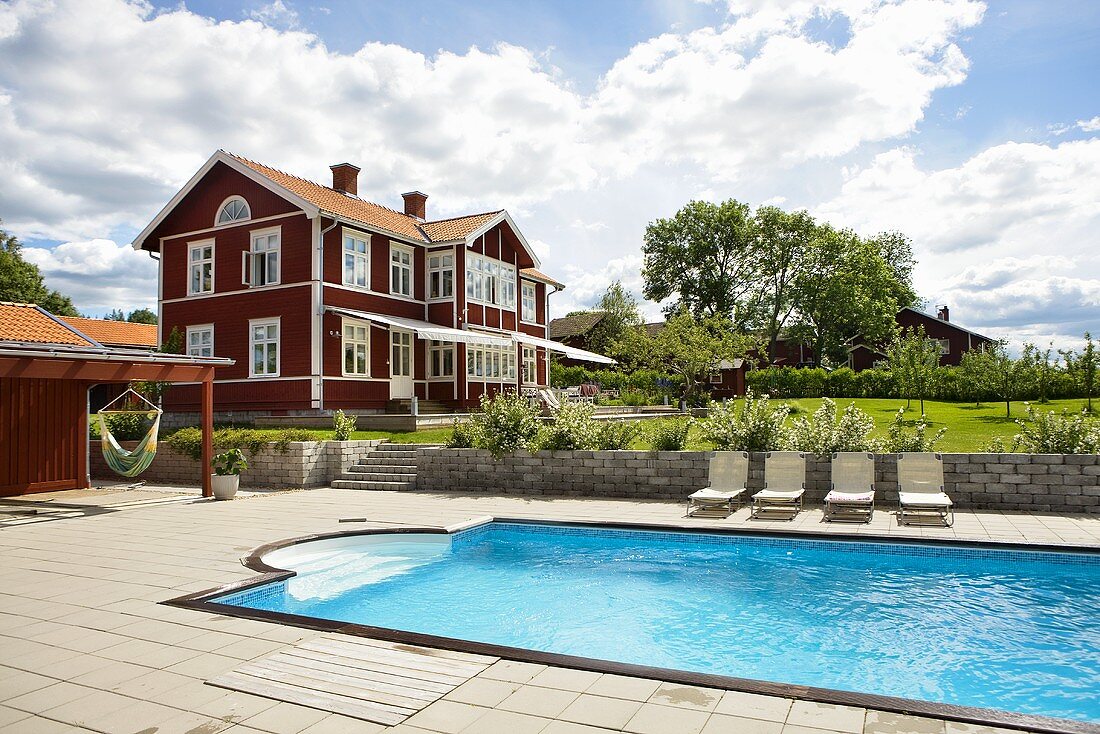 Pool mit Liegestühlen und rotbraunes Holzhaushaus mit weissen Fenstern