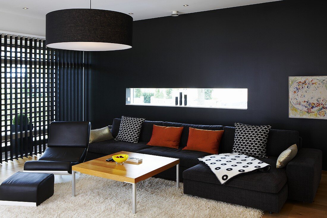 A black wall with a narrow window and a wooden lattice wall in a living room furnished with black chairs and a sofa and a wooden coffee table